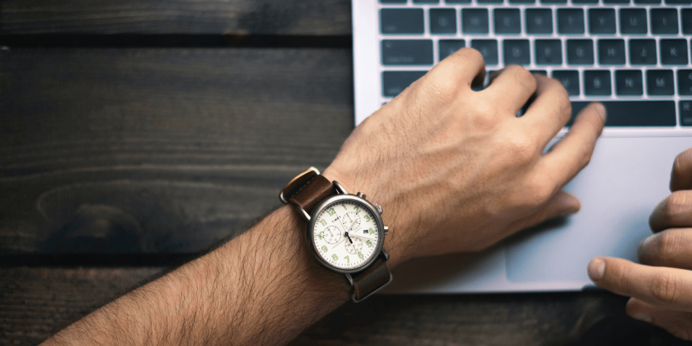 A man looking at his watch in front of a laptop