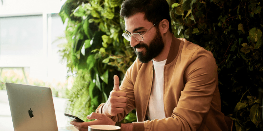 A man holding a mobile phone in front of a laptop giving a thumbs up.