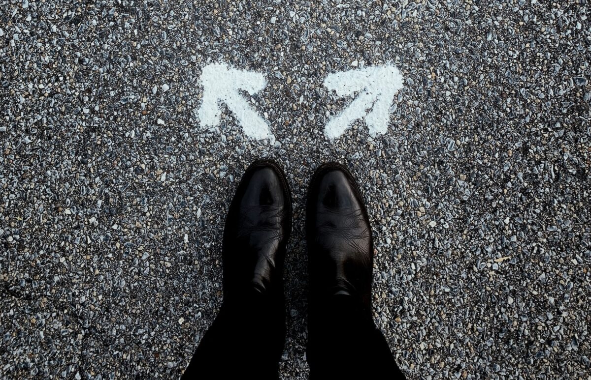 A picture of a man's feet in black dress shoes and slacks in front of arrows pointing in two different directions.